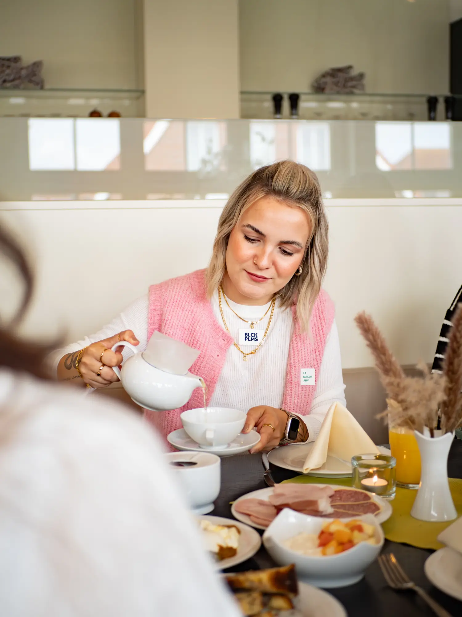 Eine blonde Frau mit kurzen Haaren sitzt im Frühstückstisch und schenkt sich einen Tee aus einer Kanne in ihre tasse ein.