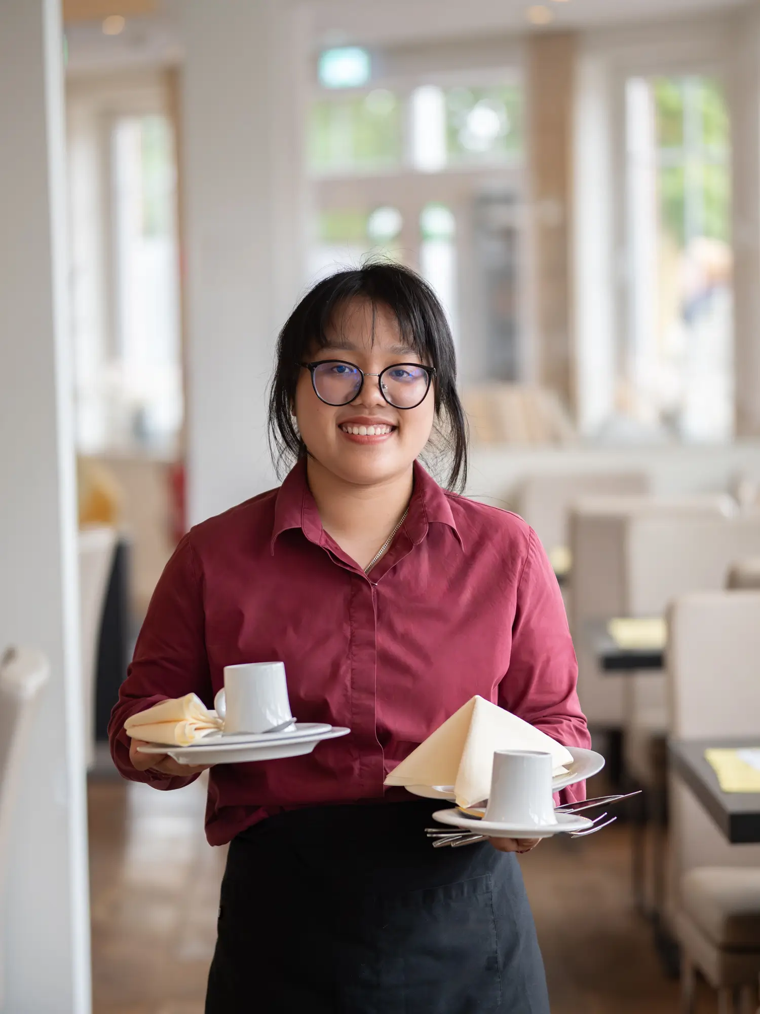 Eine weibliche Service-Mitarbeiterin blickt lächelnd in die Kamera und hält eine Tasse, einen Teller, eine Serviette und Besteck in ihren Händen.
