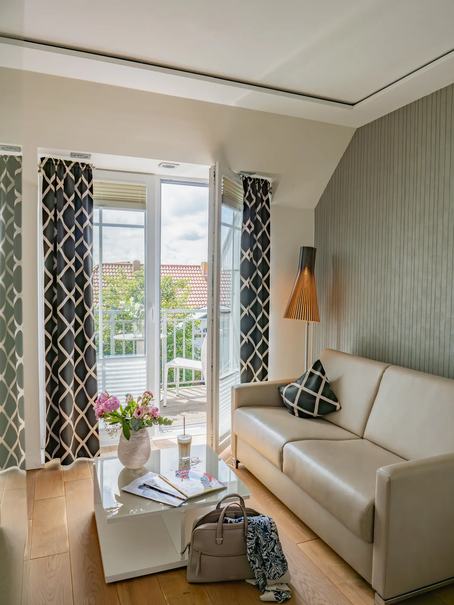 Ein Hotelzimmer mit Balkon geradeaus, Sofa rechts, davor steht ein Couchtisch. dort stehen Blumen und ein Glass Latte Macchiato.