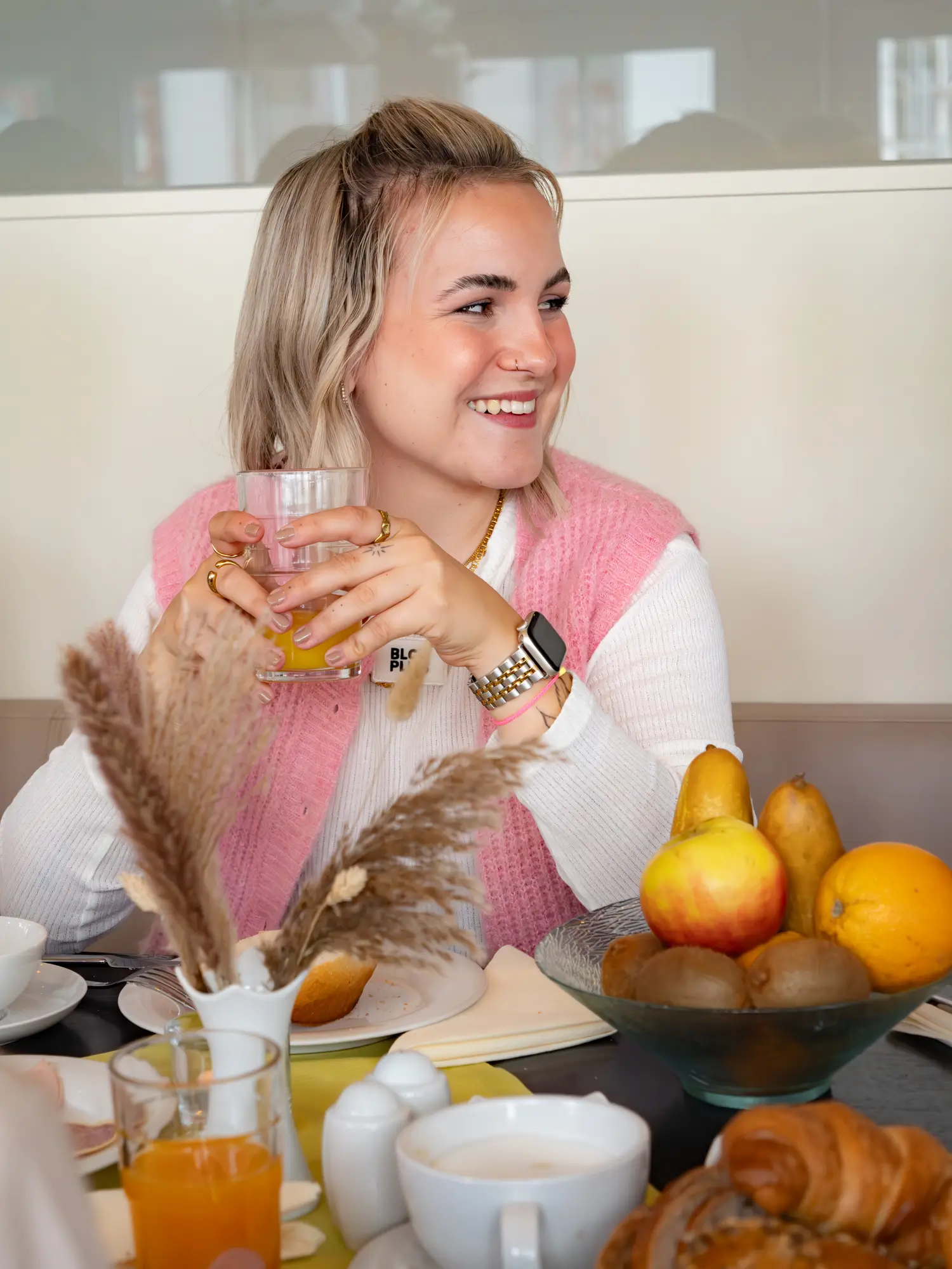 Eine blonde Frau mit kurzen Haaren sitzt am Frühstückstisch, hält eine Tasse in beiden Händen, blickt zur Seite und lächelt.
